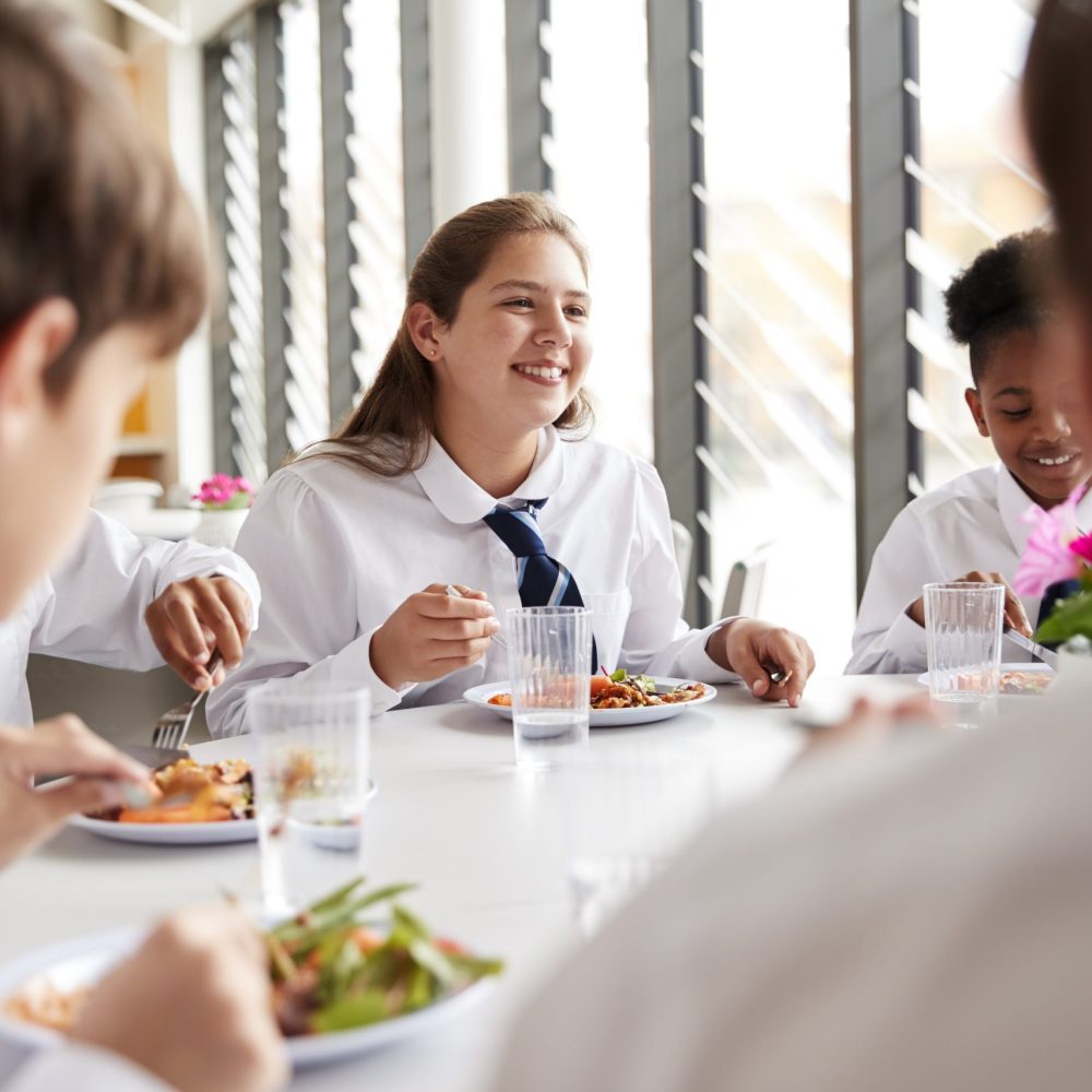 Group Of High School Students Wearing Uniform Sitting Around Table And Eating Lunch In Cafeteria